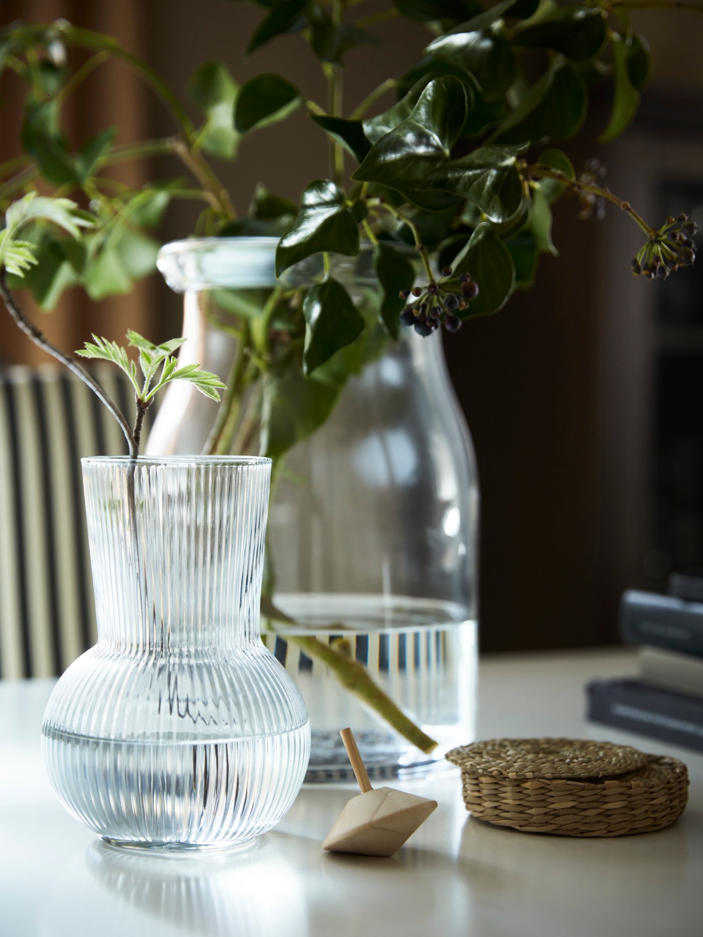 A clear glass BEGÄRLIG vase and a clear glass PÅDRAG vase, both containing decorative plant cuttings, stand on a white table.