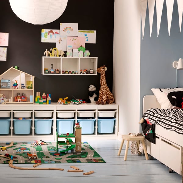 Una habitació infantil plena de joguines amb un sistema d'emmagatzematge TROFAST al llarg d'una paret negra. A la dreta, hi ha un llit SLÄKT.