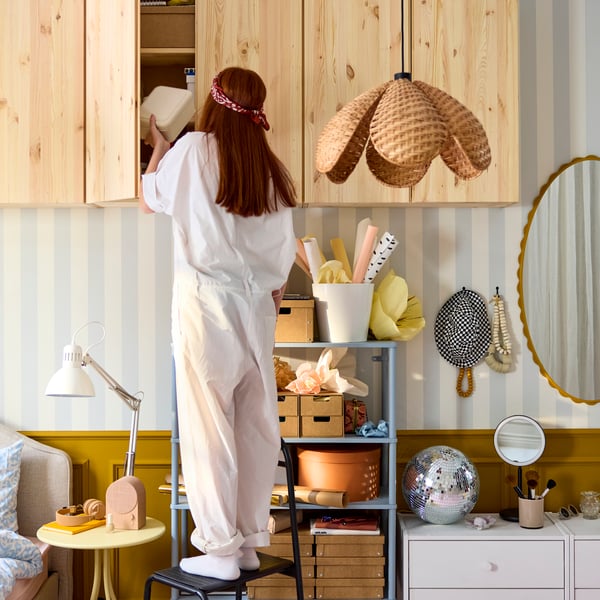 Dans une pièce lumineuse, un adolescent monte sur un tabouret pour atteindre le haut d'une armoire IVAR en pin naturel fixée au mur.