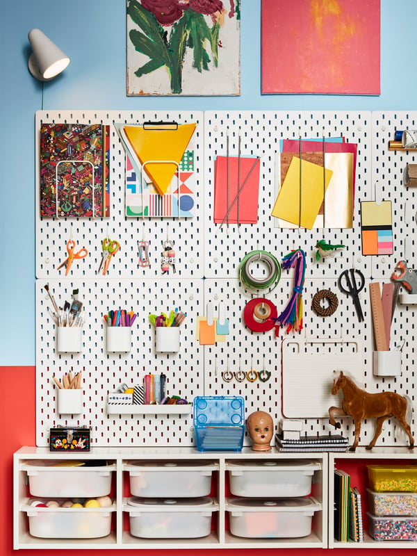 White SKÅDIS pegboards with accessories such as containers and hooks holding artist’s supplies are attached to a wall.