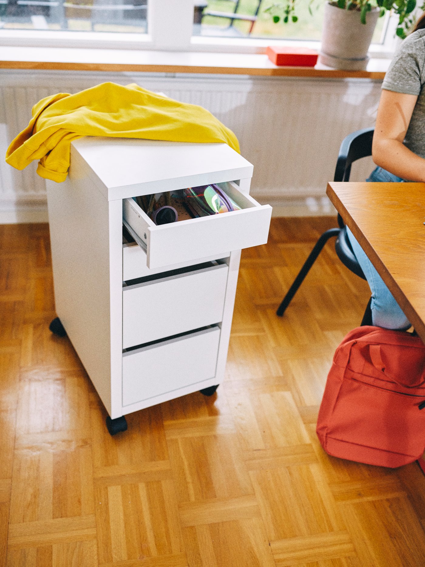 A white MICKE drawer unit with a yellow jacket on top, beside a person sitting at a table with a STARTTID backpack.
