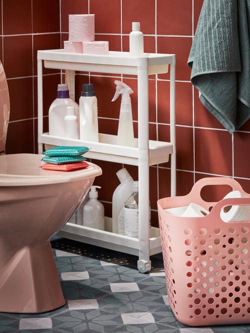 Ein weißer VESKEN Rollwagen mit Reinigungsmitteln und Badezimmerutensilien, der in einer Ecke neben einer Toilette steht.