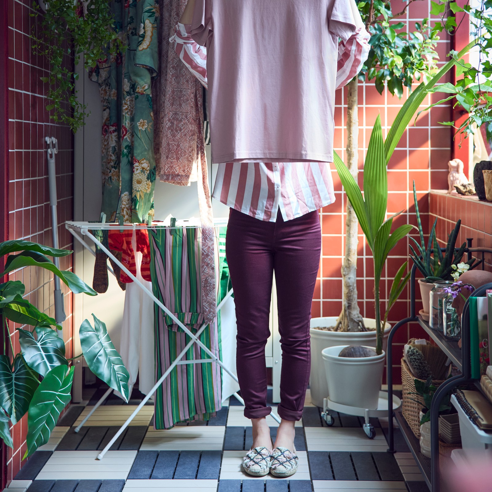 A white JÄLL drying rack holding clothes is in a tiled, enclosed balcony with greenery where a person is hanging up a shirt.