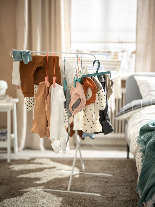 Clothes on SLIBB hanging pegs and a SLIBB hanger, arranged on a white MULIG drying rack next to a bed.