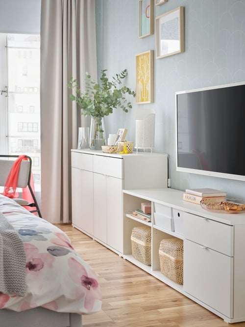 Ein weißes VIHALS Sideboard und eine TV-Bank mit einem Fernseher darüber an einer Wand am Ende eines GLADSTAD Bettgestells. Dahinter sind Bilder zu sehen.