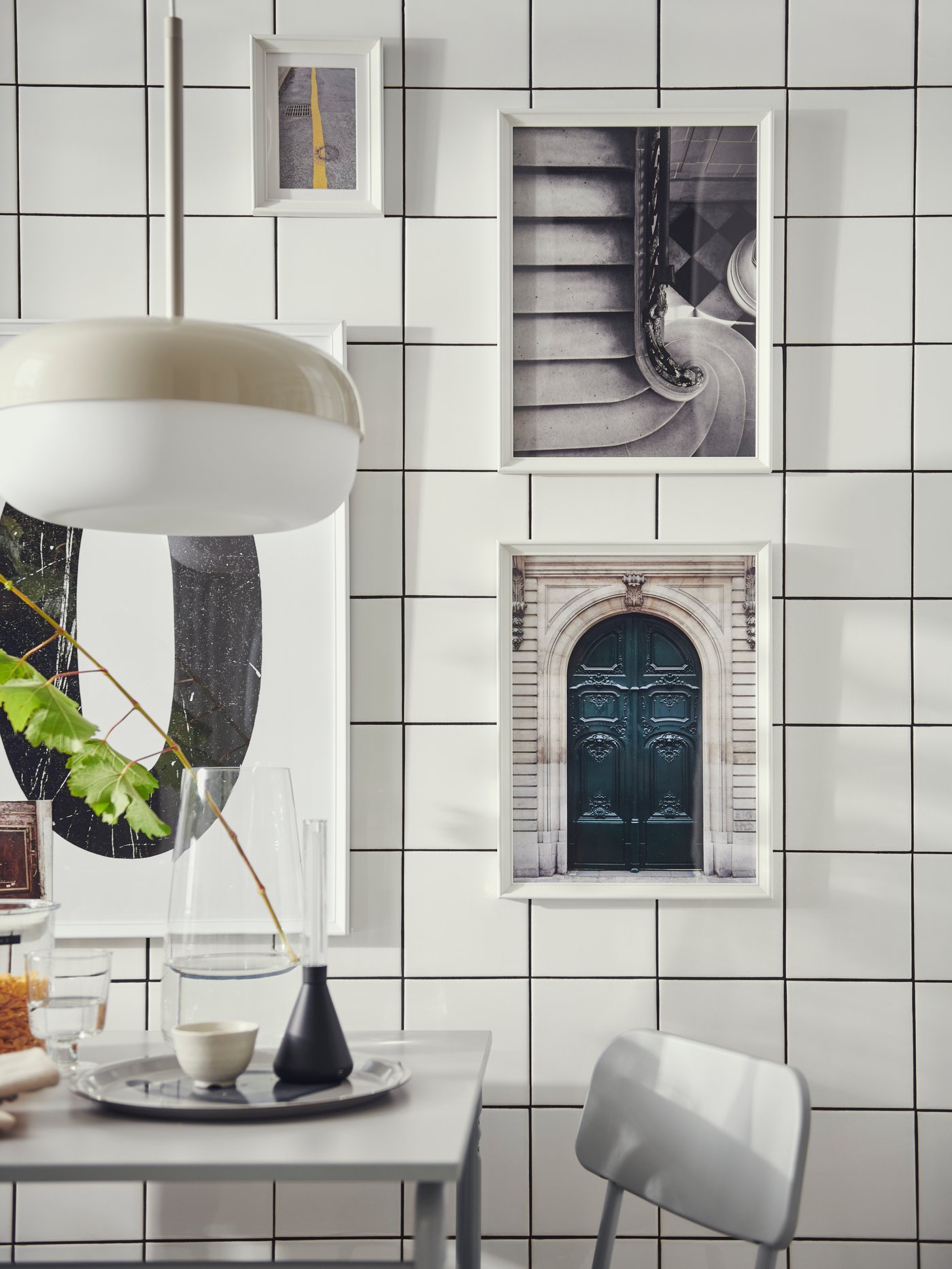 Cocina con cuadros en marcos KNOPPÄNG colocados en una pared de azulejos. Hay una mesa GRÅSALA bajo una lámpara de techo BLÅSVERK.