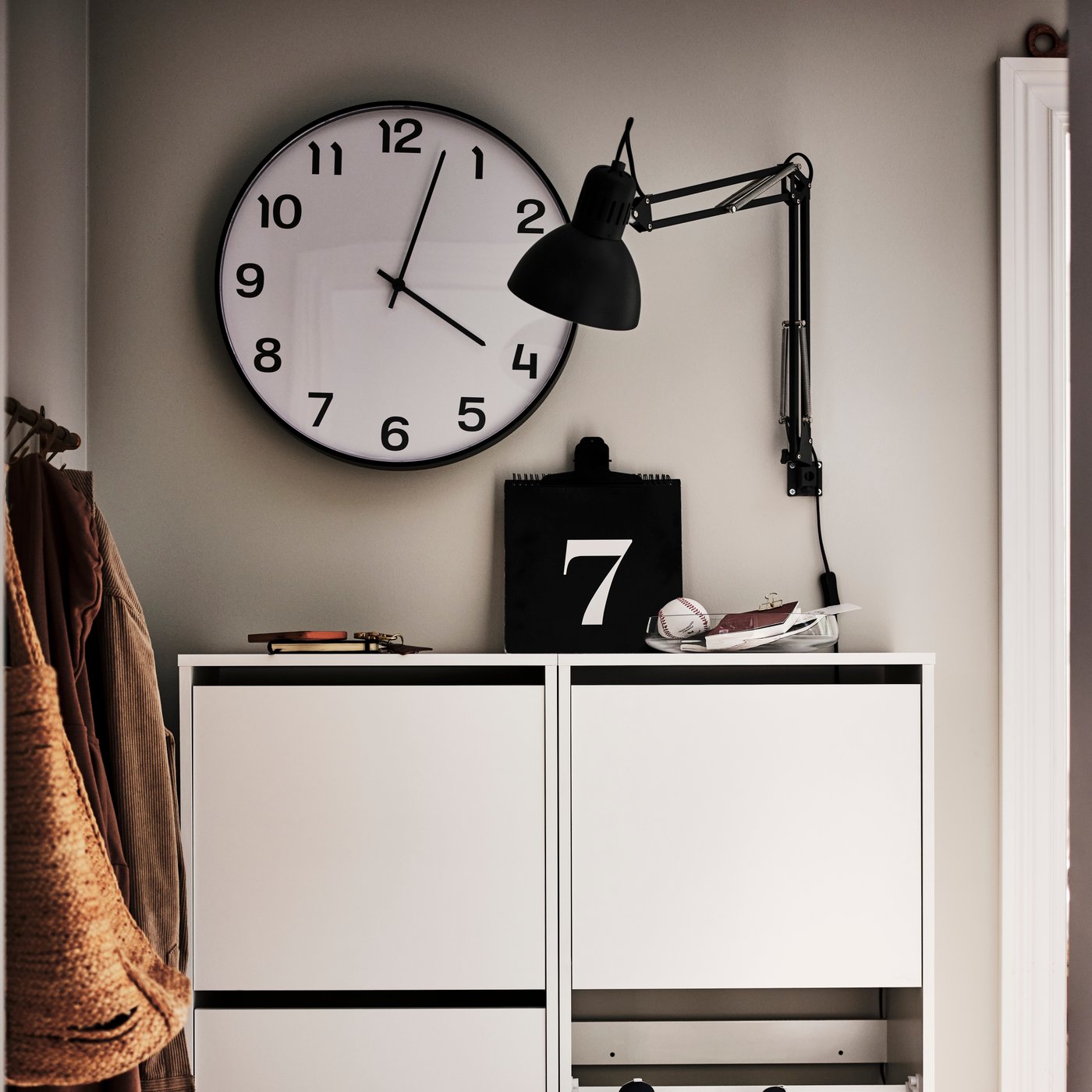 A black PLUTTIS wall clock hangs on a wall above a white storage unit. 
