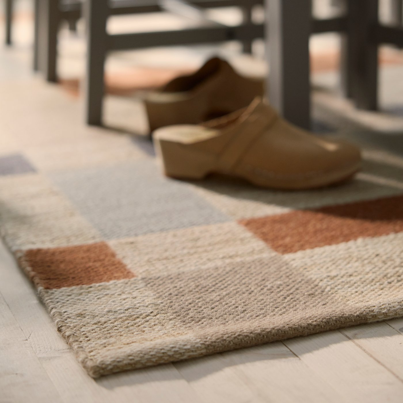 Tapis tissé plat BLÅLJUS dans des teintes naturelles grises et brunes sous des chaises en bois, sur un sol en bois clair.