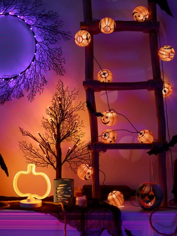 Small decorative wooden ladder sitting on mantle. Room dimly lit by KUSTFYRLED lights. Small KUSTFYR pumpkin string lights wrapped around ladder.