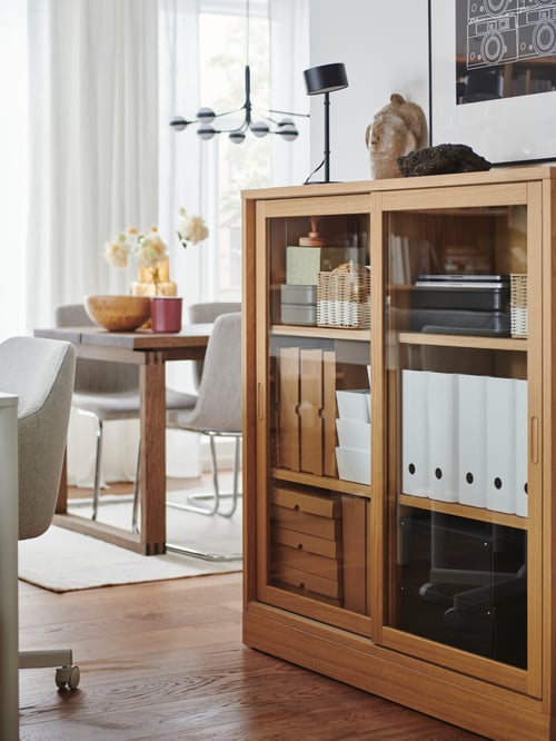 A living room showing a TONSTAD cabinet with sliding glass doors filled with baskets, and magazine files, beside a chair.