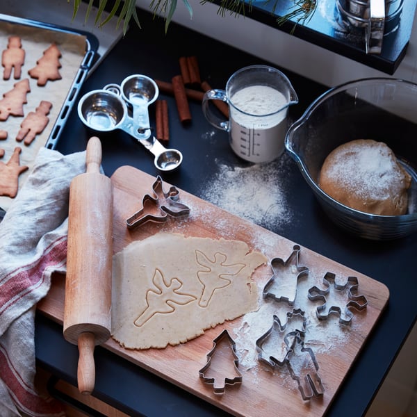 Una tabla de madera PROPPMÄTT, moldes de galletas VÄRMER, un uslero de madera MAGASIN, recipientes de vidrio VARDAGEN con masa y harina sobre una encimera oscura, en medio de una preparación de galletas. 