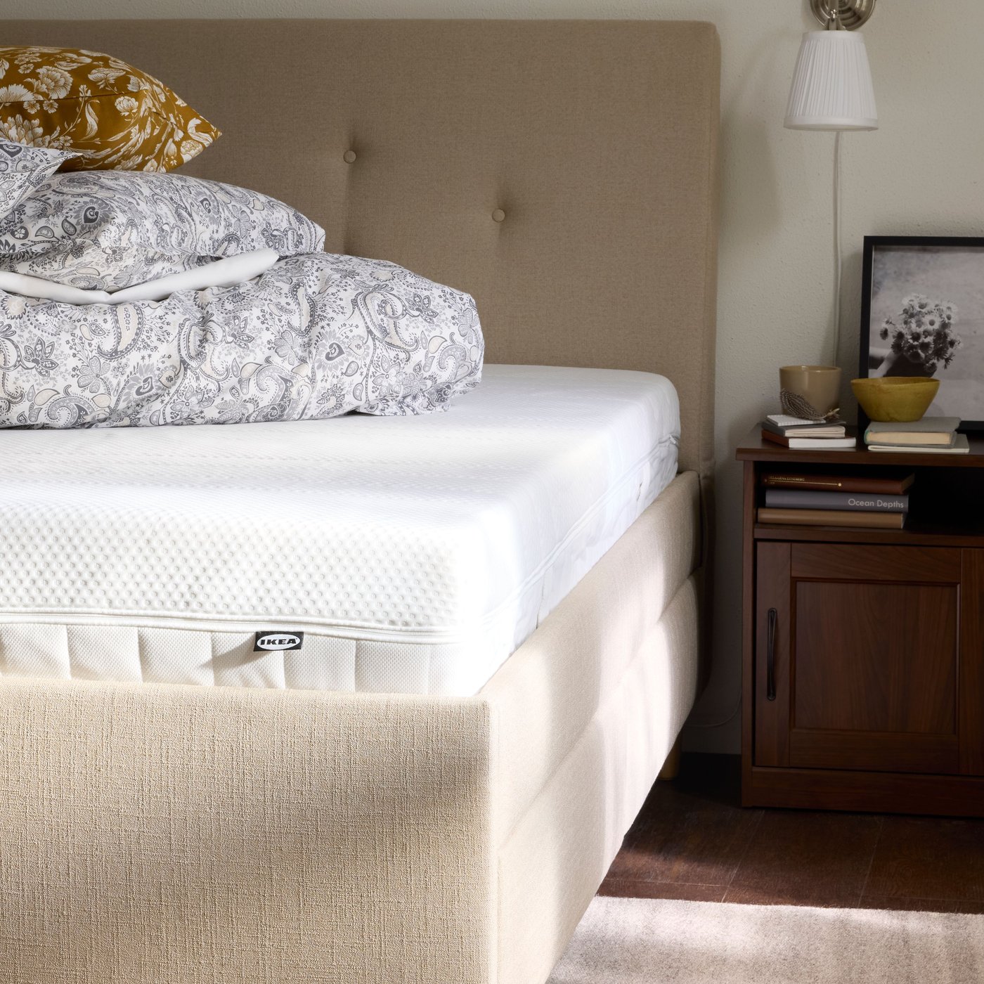 A made bed with a beige headboard and a white mattress stands next to a bedside table with books, a bowl, and a lamp in a calm bedroom.