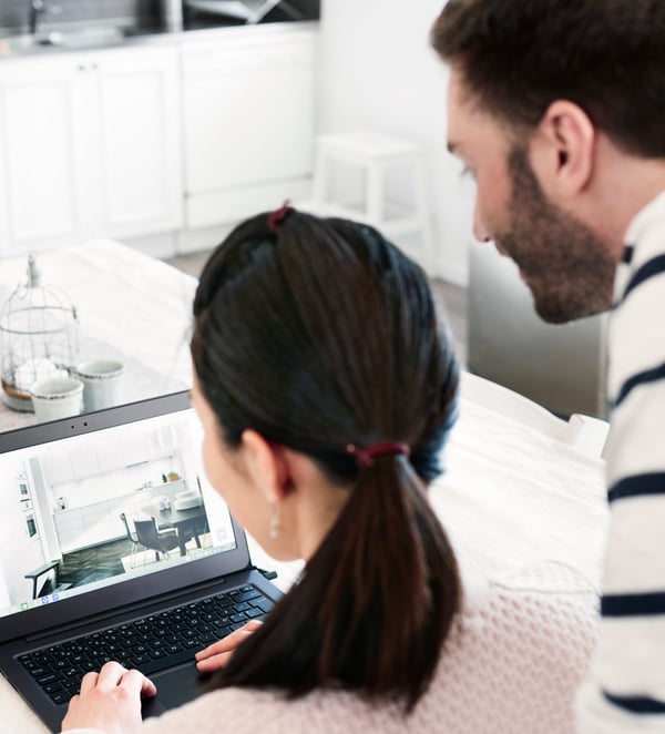 A couple is looking at an open laptop where there is the plan of a white kitchen.