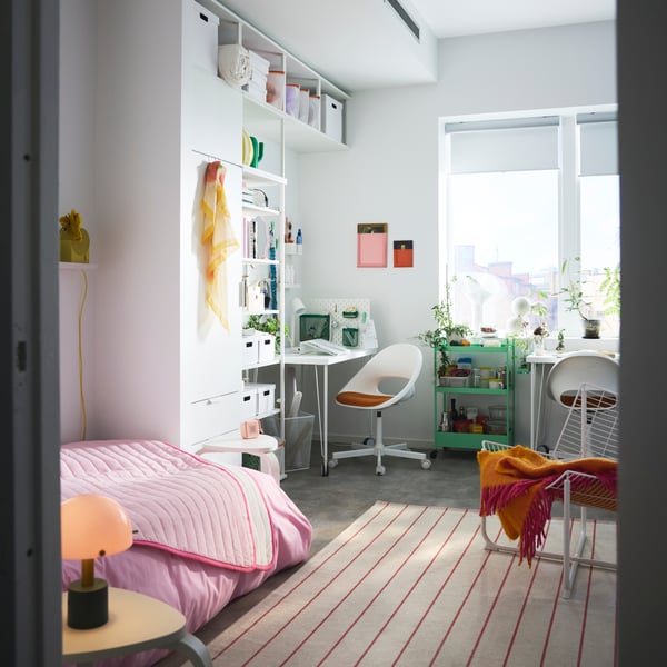 A shared college dorm showing a bed with a pink bedspread, a wardrobe with open shelves, a desk, desk chair and trolley.