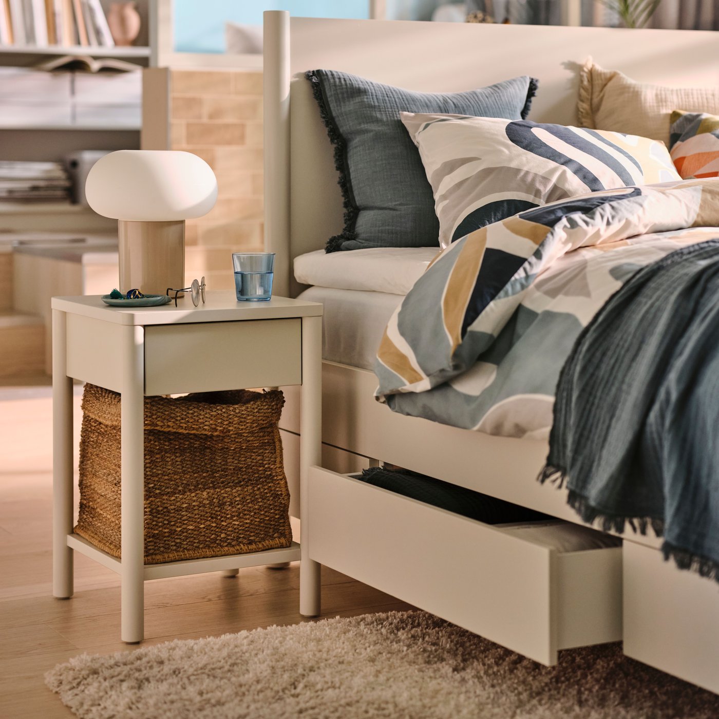 An off-white TONSTAD bedside table beside an off-white TONSTAD bed covered in MYGGLASVINGE multicolour bed linen.