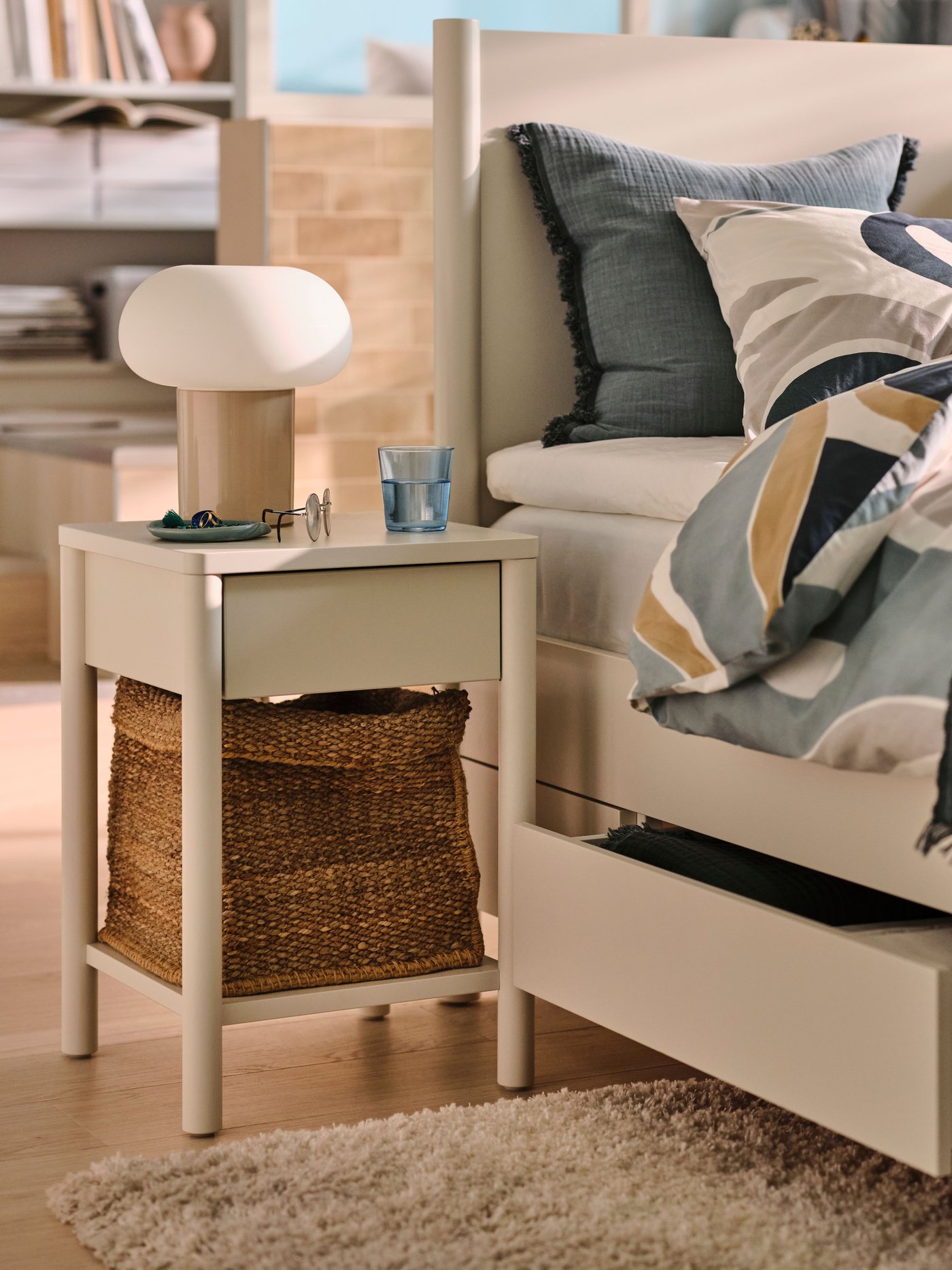An off-white TONSTAD bedside table beside an off-white TONSTAD bed covered in MYGGLASVINGE multicolour bed linen.