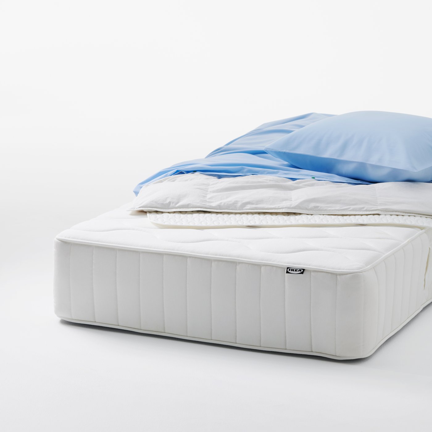 An uncovered VÅGSTRANDA pocket sprung mattress with light bedding placed on top in a clean studio setting.