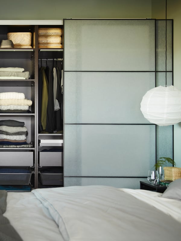 A bedroom with an open PAX/SVARTISDAL wardrobe full of clothes beside a double bed with a REGOLIT lamp hanging overhead.