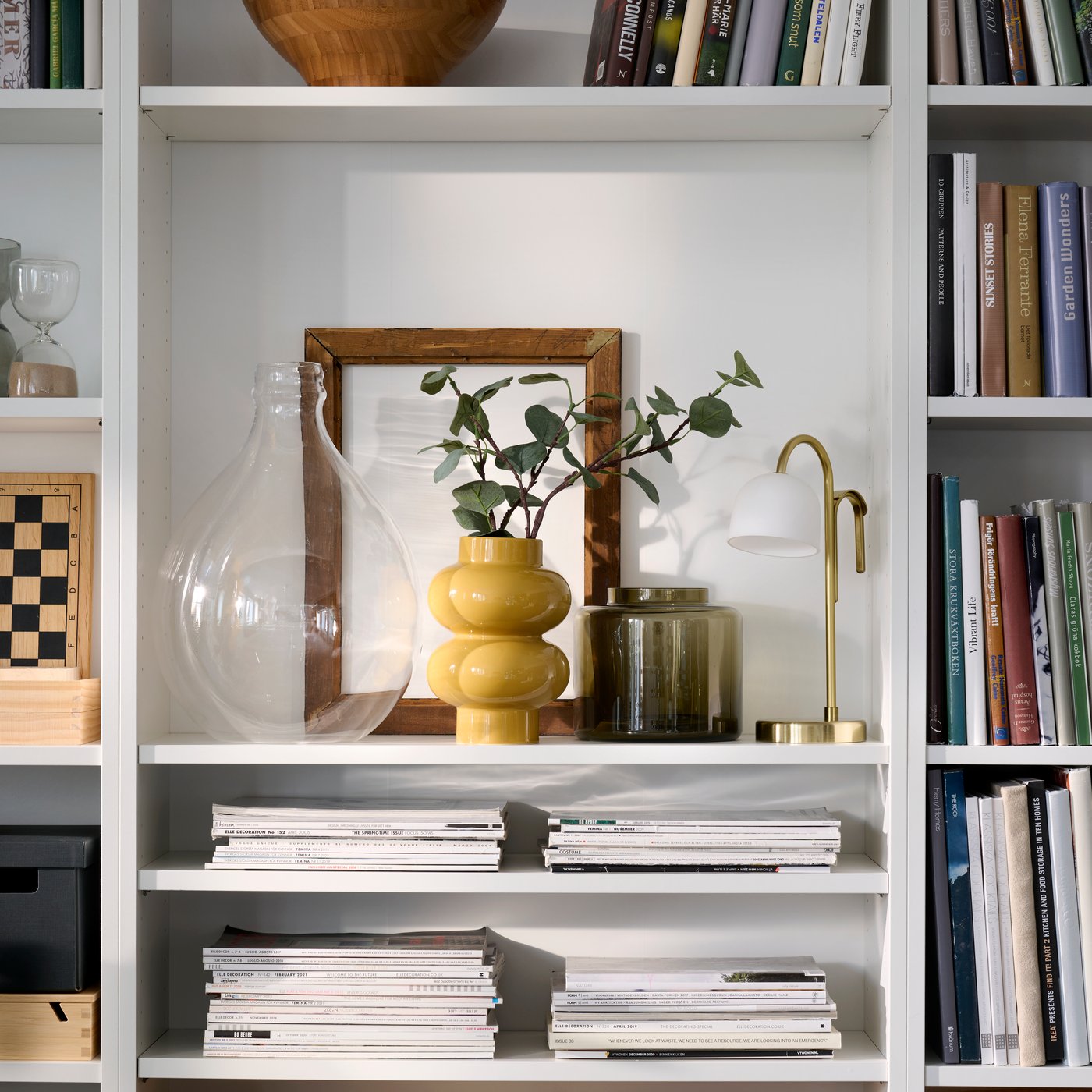 Prydligt organiserade vita BILLY bokhyllor med en gul-beige PÄRLHÄGG-vas och en TVÅMASTAD portabel lampa som delar ett hyllplan.