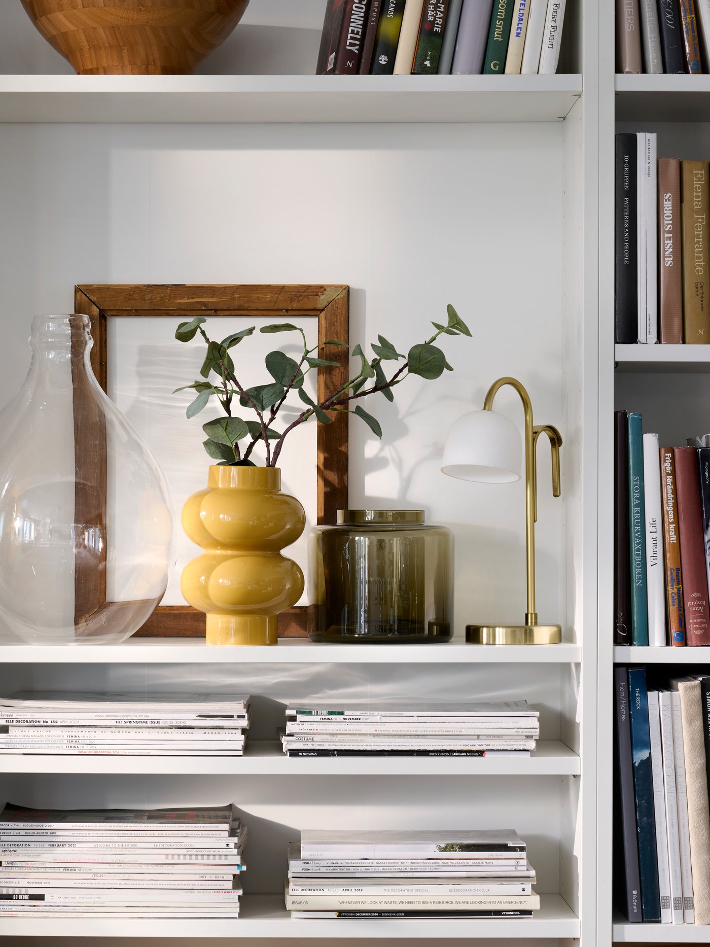 Neatly organised white BILLY bookcases with a yellow-beige PÄRLHÄGG vase and a TVÅMASTAD portable lamp sharing a shelf.