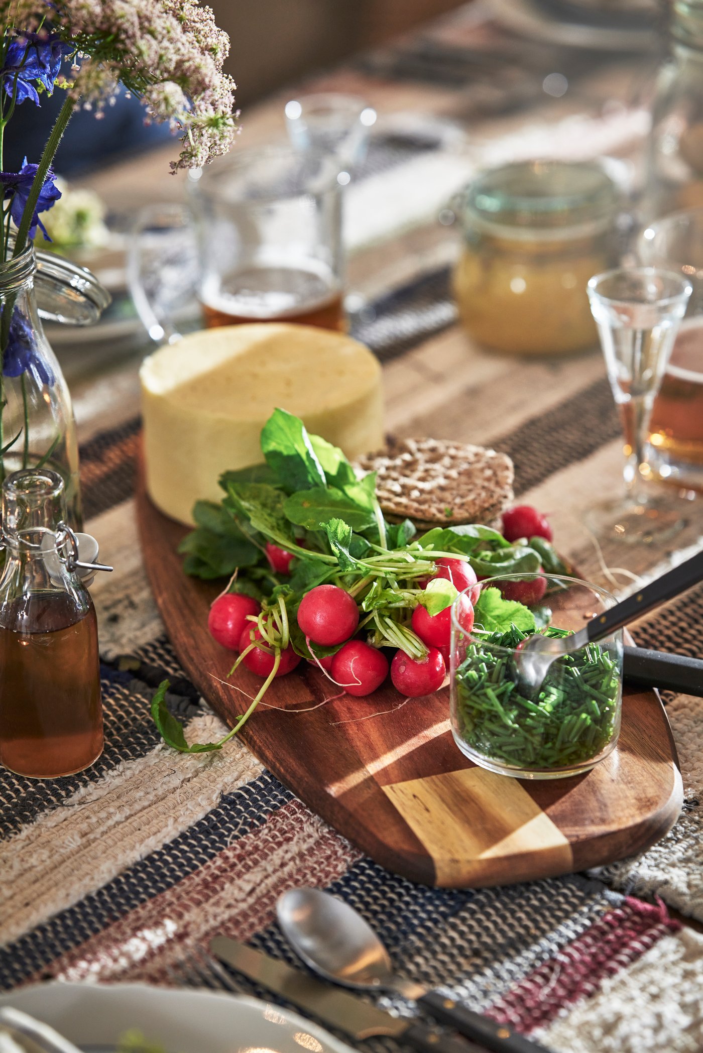 Närbild av ett dukat bord med en träbricka som rymmer en rund ost, färska röda rädisor med gröna blad och knäckebröd; runtom syns glas, små flaskor med dryck, en sked, textilier i naturtoner och mjukt dagsljus.