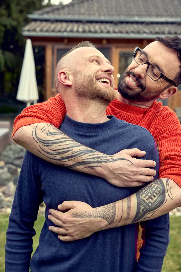 Daniel hugs his partner Martin from behind, standing in front of their home.