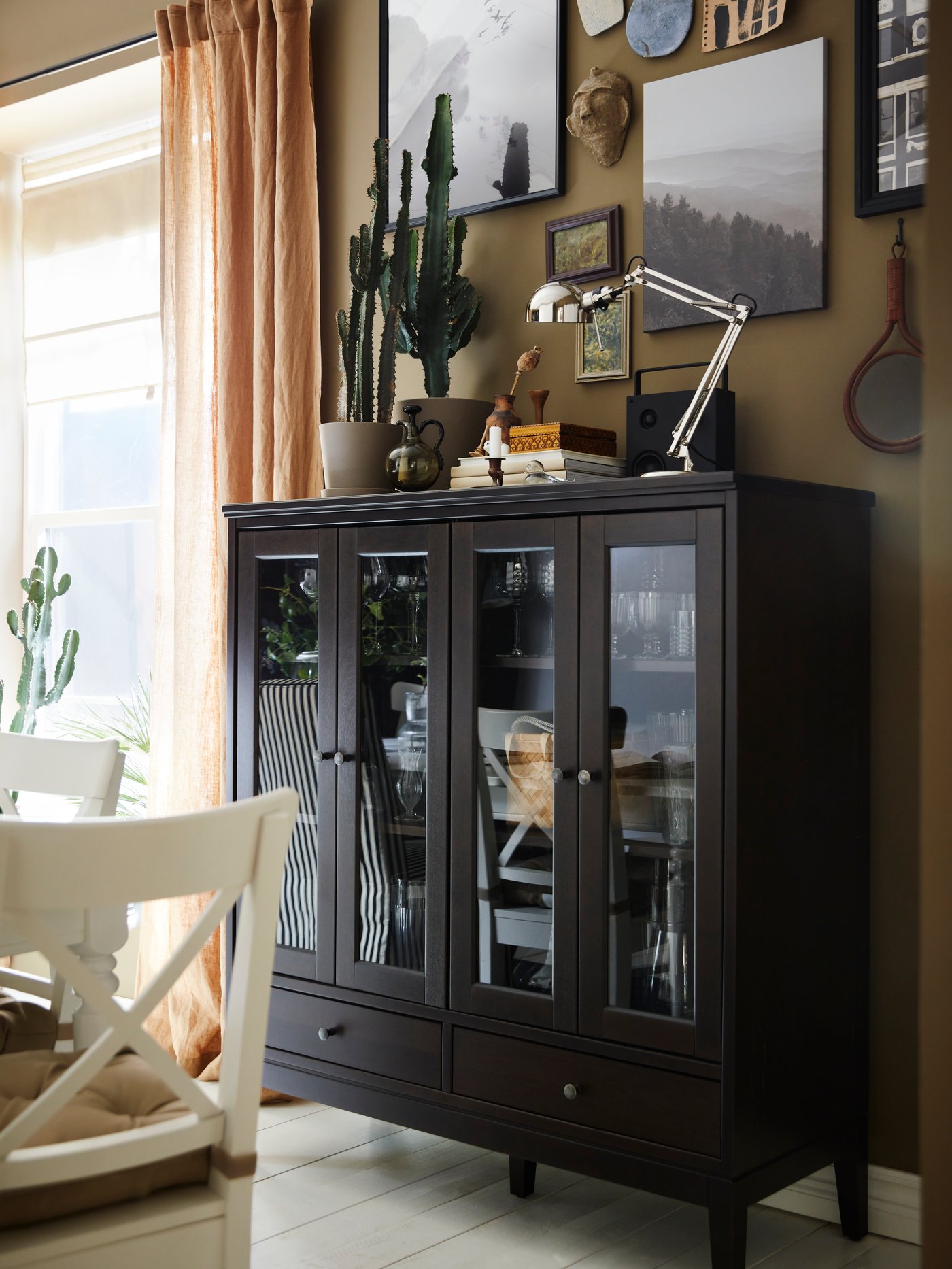 Varios objetos, entre ellos una lámpara de trabajo FORSÅ y algunas plantas, están encima de un gabinete IDANÄS con puertas de vidrio.