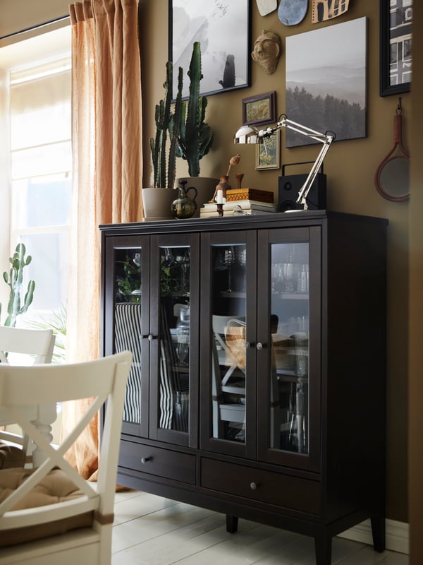 Various objects including a FORSÅ work lamp and some plants stand on top of an IDANÄS cabinet with bi-folded glass doors.
