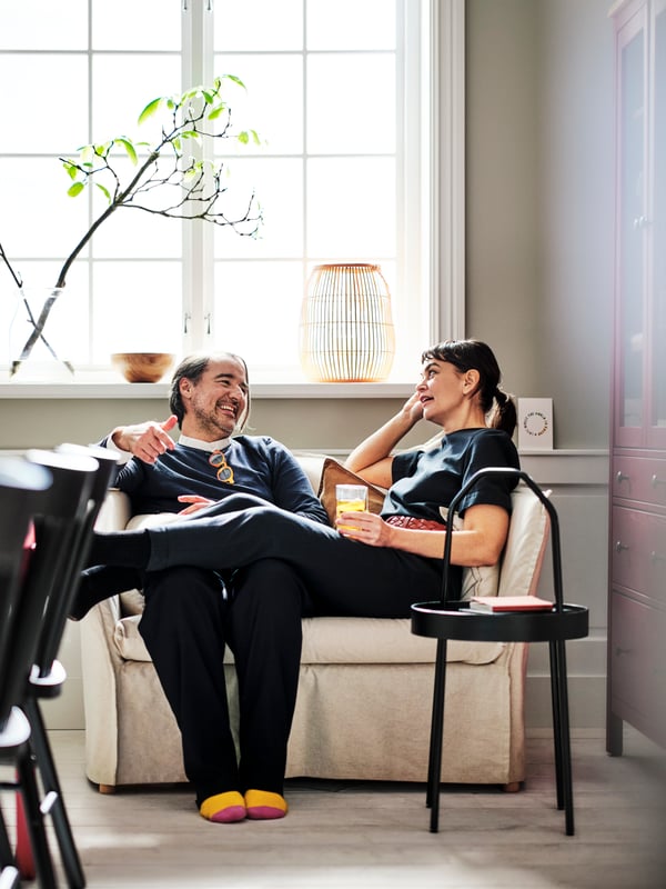 Two people chatting while seated on a natural BACKSÄLEN 1,5-seat armchair near a black BURVIK side table.