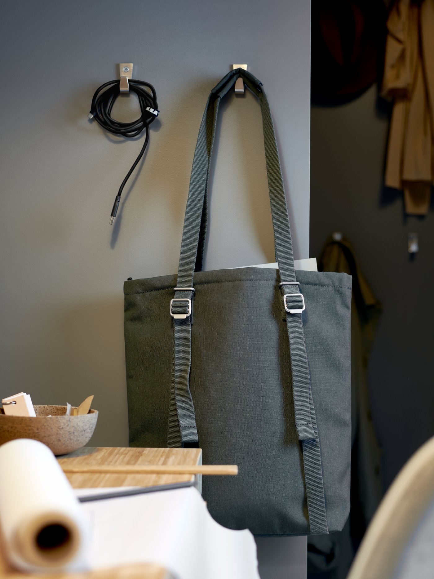 An olive-green DRÖMSÄCK tote bag hanging on a hook on a wall, next to another hook with a wound phone-charger cable on it.