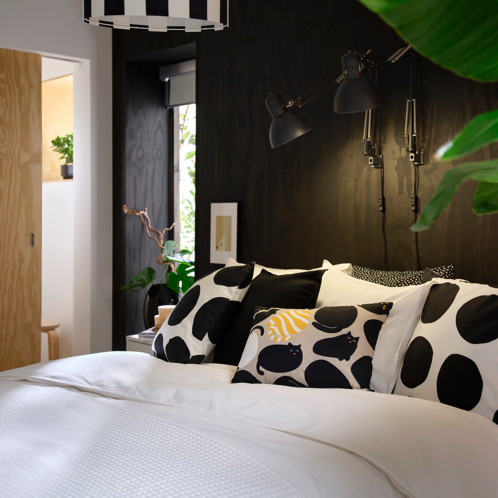 ÄNGSLILJA duvet cover in white washed cotton on a bed with patterned cushions in a bedroom with dark wood wall.