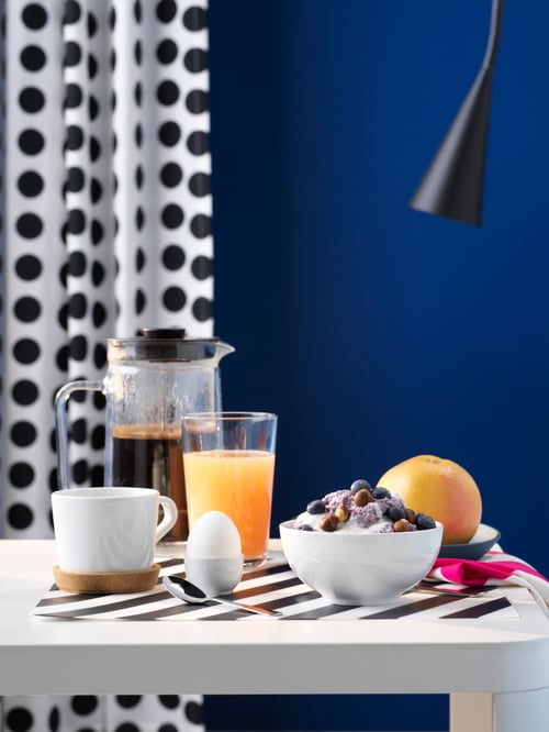 Cameră de zi cu cafetieră, un pahar cu suc de portocale, bol cu fructe, o cană și un ou așezate pe o masă albă.