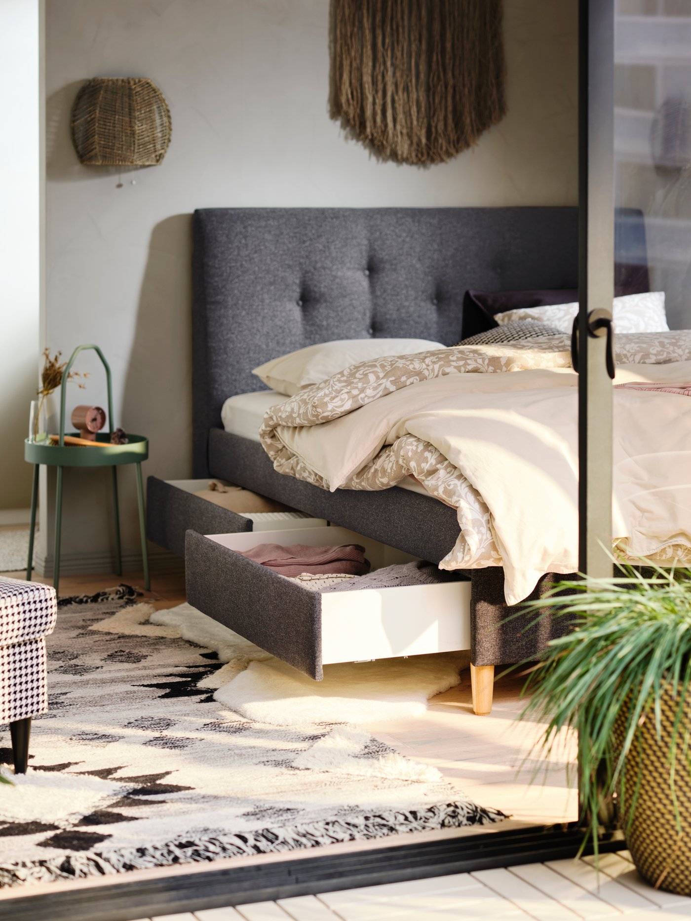 Una cama tapizada IDANÄS en gris con ropa de cama de flores y lisa con cajones de almacenaje abiertos donde se guardan textiles doblados.