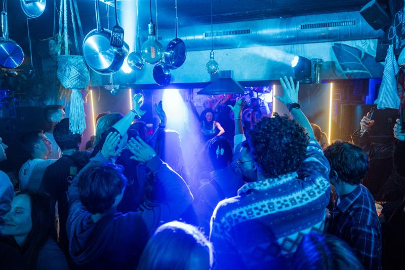 Menschen feiern ausgelassen in einem Club mit blauer und violetter Beleuchtung; einige heben die Hände im Takt der Musik, während ein DJ auf der Bühne performt. Die Decke ist mit Lampen und Pflanzen dekoriert.