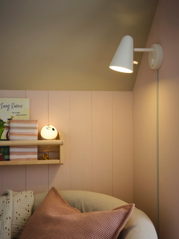 A FLISAT wall storage holds books and a KORNSNÖ LED night light on a wall above a TULLSTA chair with a pink cushion on it.