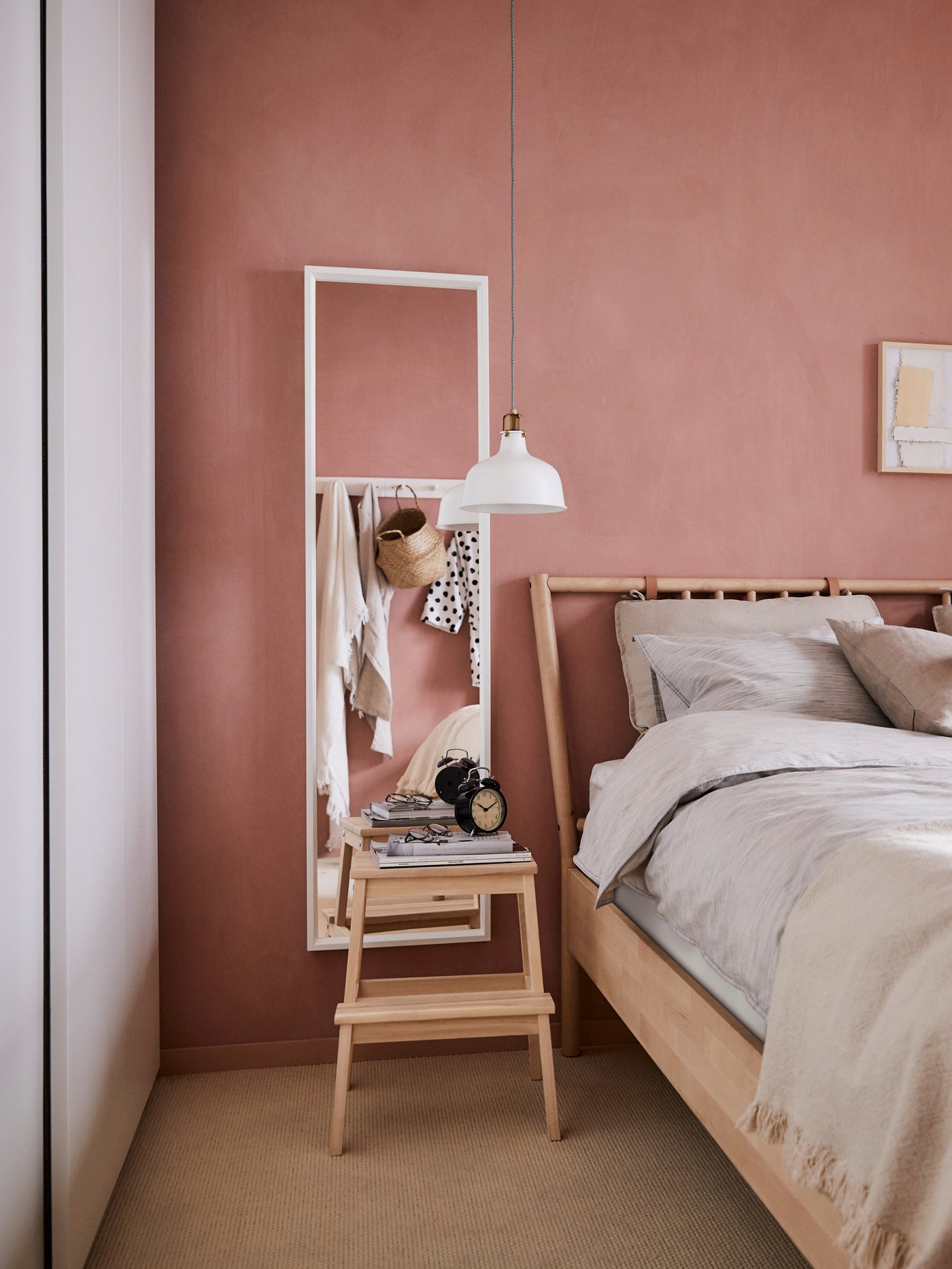 Mirall NISSEDAL amb marc de fusta blanc en una paret d'un to rosat amb un tamboret de fusta al davant al costat d'un llit.