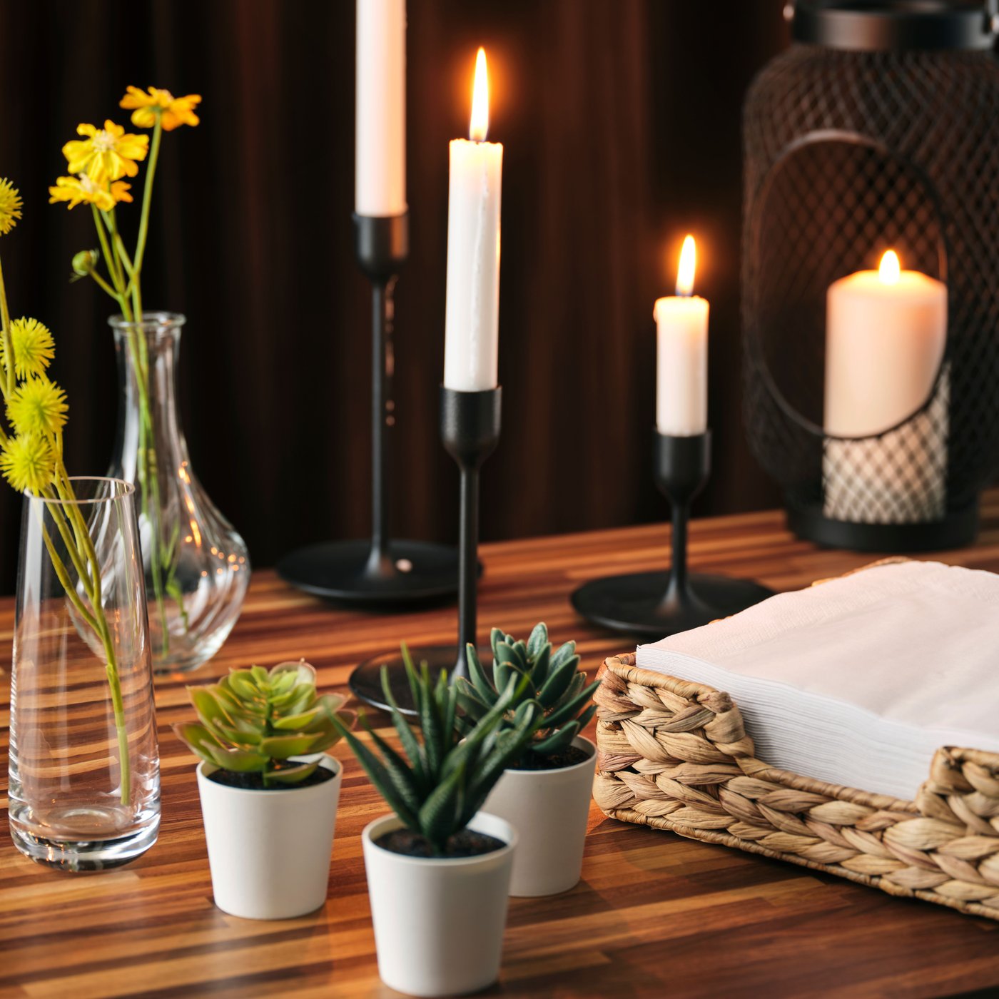 A black FULLTALIG candlestick set of three holding tall white candles, accented by small potted succulents and flowers.