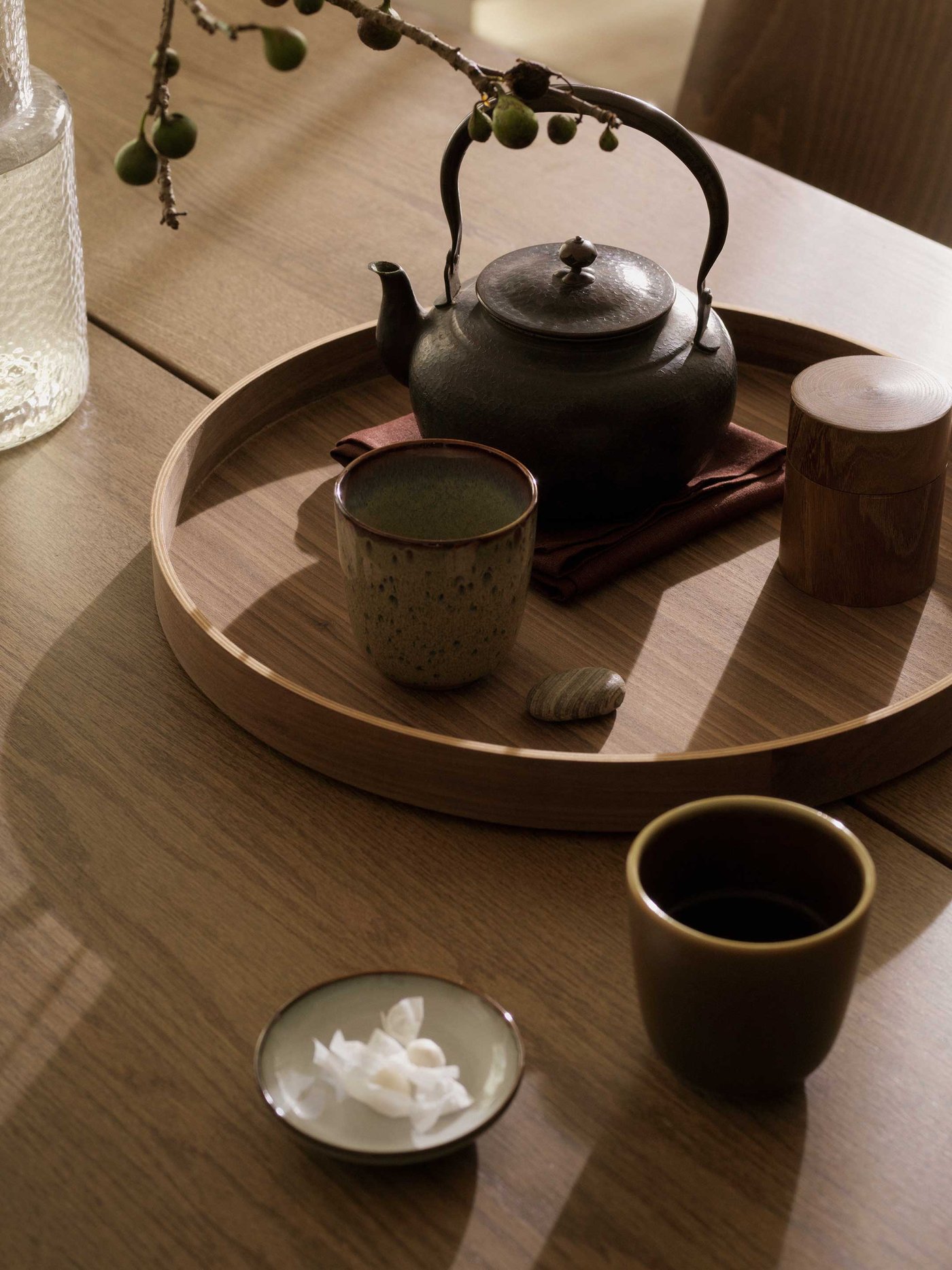A delightful close-up of a wooden dining table, set for a tranquil tea moment. A round wooden tray holds a rustic, dark cast-iron teapot, a speckled ceramic cup, and a charming wooden container, all resting on a deep red napkin. A small grey pebble adds a touch of natural calm to the arrangement. Next to the tray, a clear glass bottle with a branch adorned with green berries brings a fresh, organic feel to the setting. Another dark brown cup and a small dish with delicate white petals or paper are also on the table, inviting you to slow down and enjoy the simple pleasures.