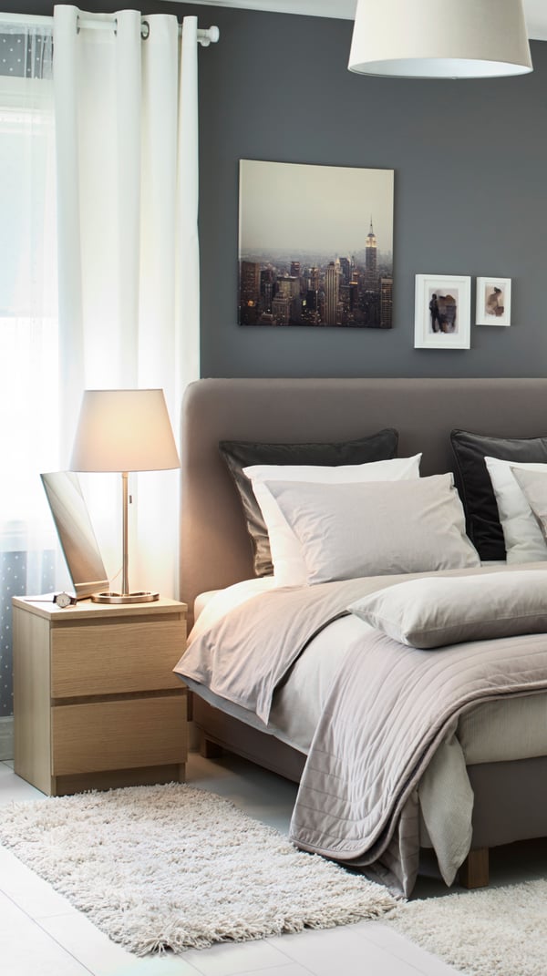 A grey FINNSNES divan bed with bed linen including light brown-red ÄNGSLILJA pillowcases next to a GLADOM tray table.