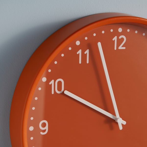 A close-up of an orange BONDIS clock hanging on a light-blue wall. The white minute and hour hands display the time.