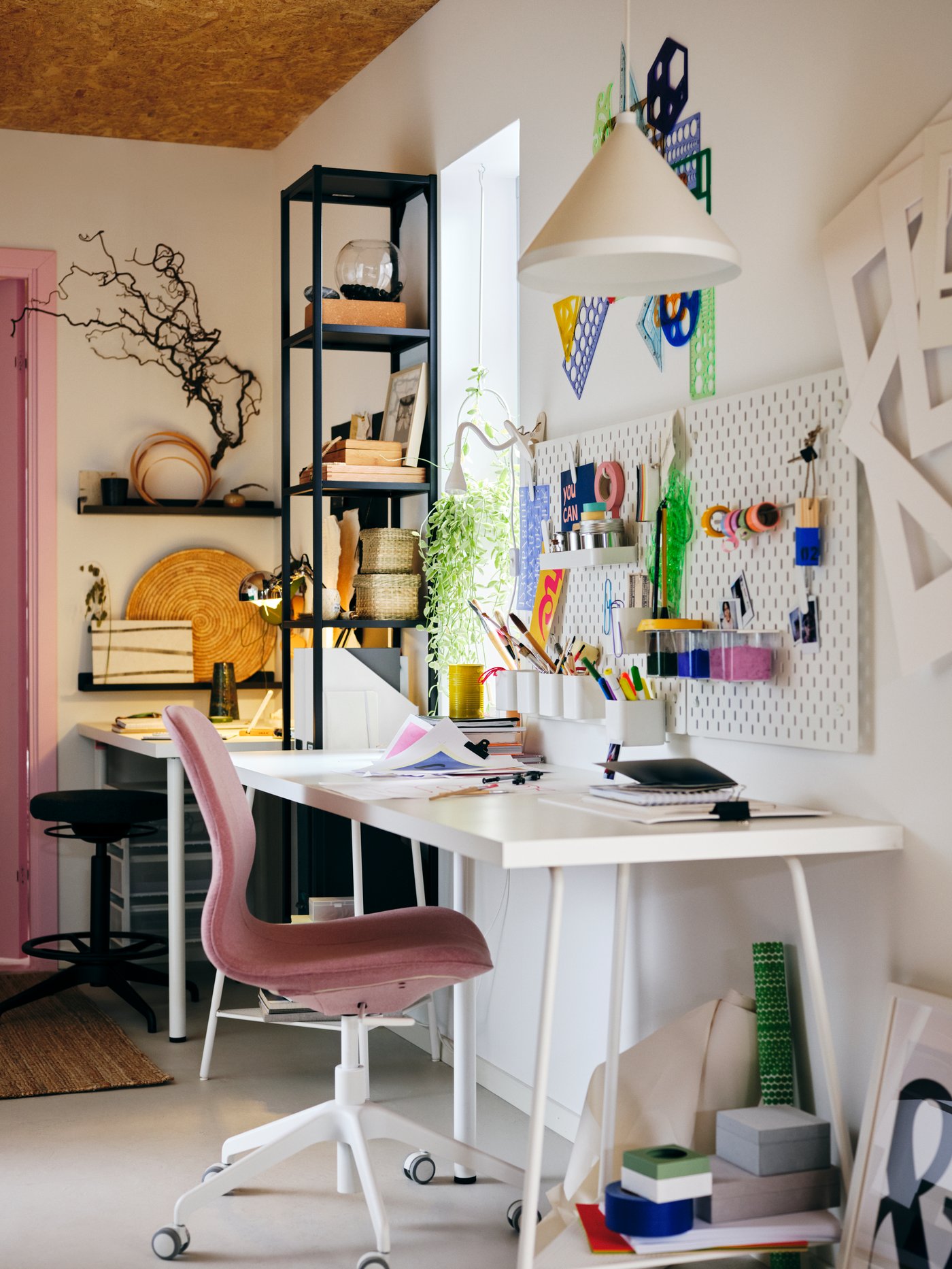 Espacio de trabajo con un escritorio, una silla de reuniones rosa, una lámpara de techo blanca, una estructura alta con estantes y una combinación de tablero.
