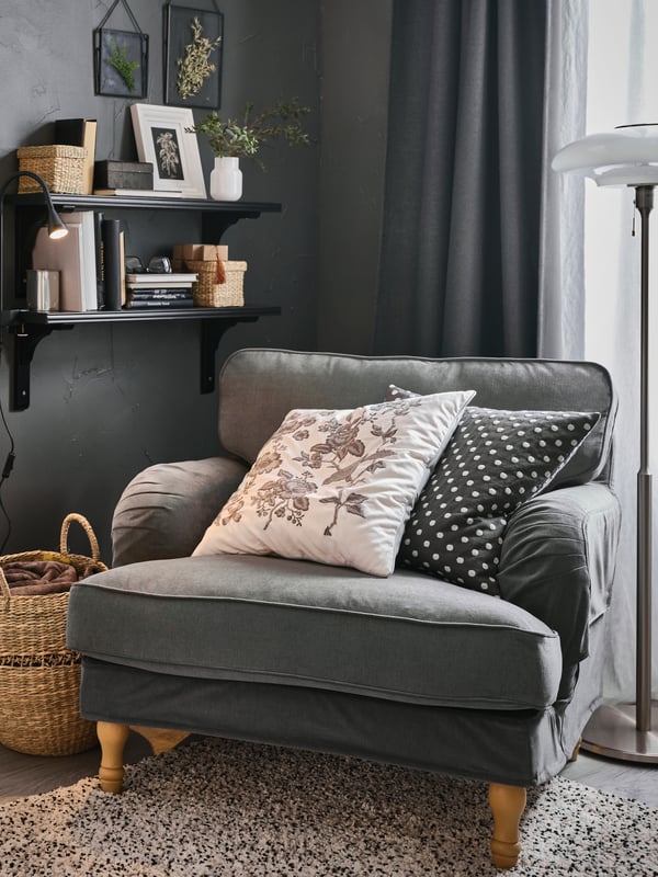 A STOCKSUND armchair with two extra cushions stands in a corner, near two brown-black BERGSHULT shelves on a wall.