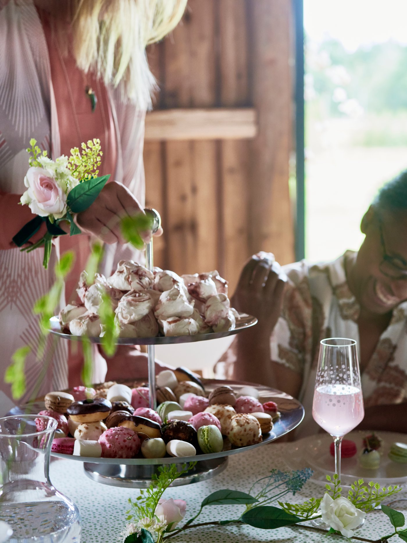 A woman holds an INBJUDEN serving stand and offers a selection of sweets to a guest at wedding in a rustic setting.
