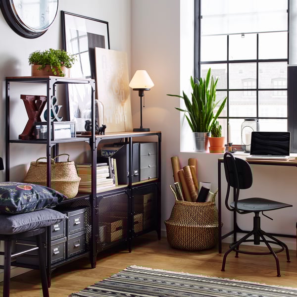An industrial-style living room with a black metal open shelving unit featuring paintings, a table lamp, and a black office chair.