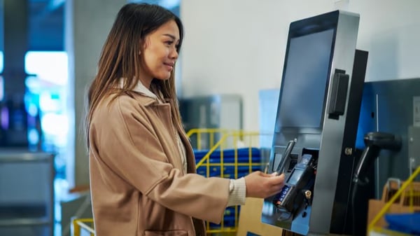 Una donna con una giacca beige effettua un pagamento contactless con il suo smartphone a un terminale.
