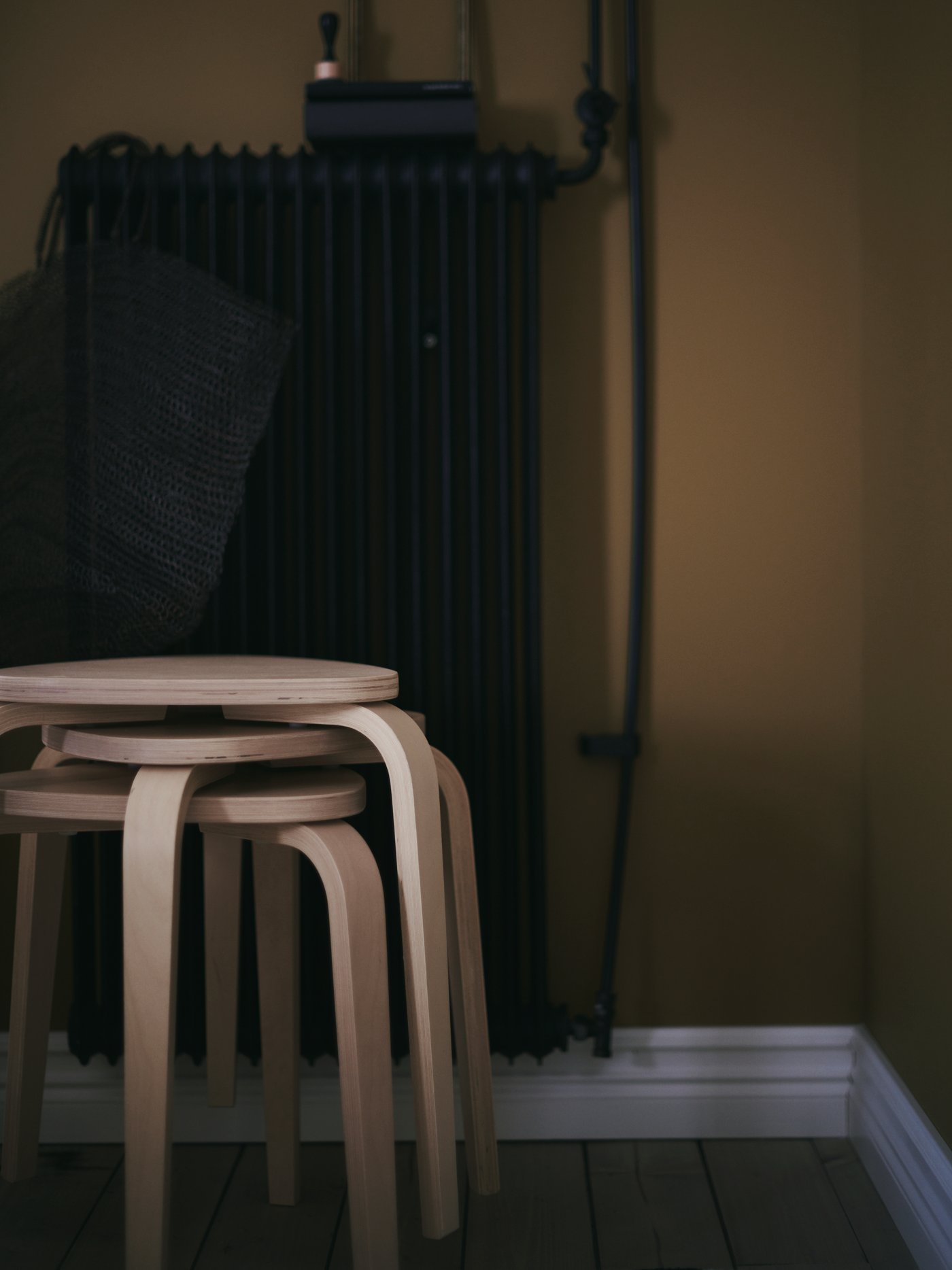 A stack of KYRRE stools in birch, in front of a dark coloured metal radiator with a bag hanging from it.