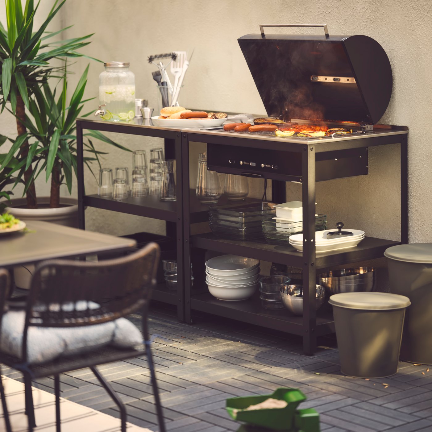 Ein Griller steht an einer Wand und auf dem Griller wird Fleisch gegrillt. Daneben stehen 2 Stühle und 1 Tisch.