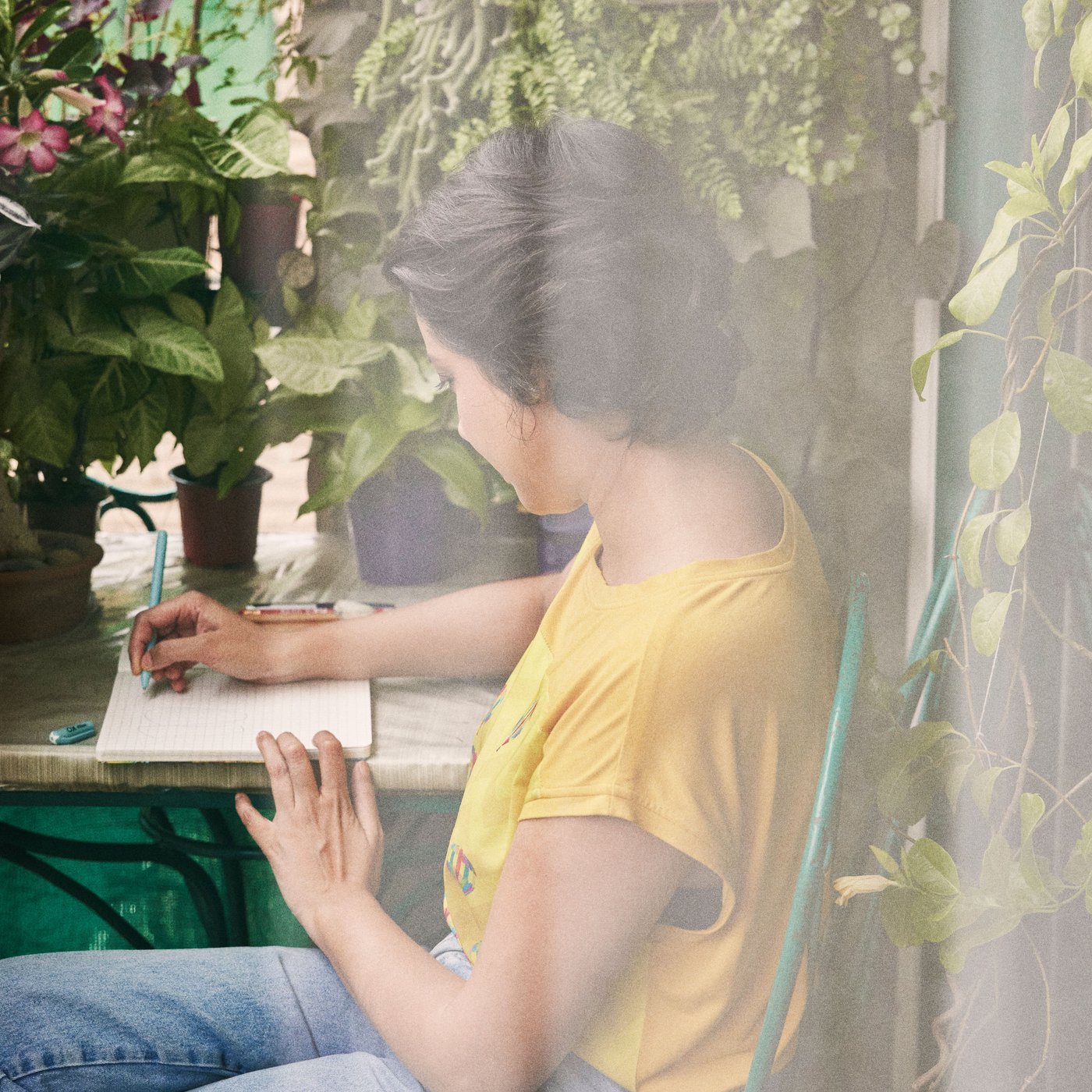 A woman in a yellow shirt sits at a table in her garden and writes in her book.