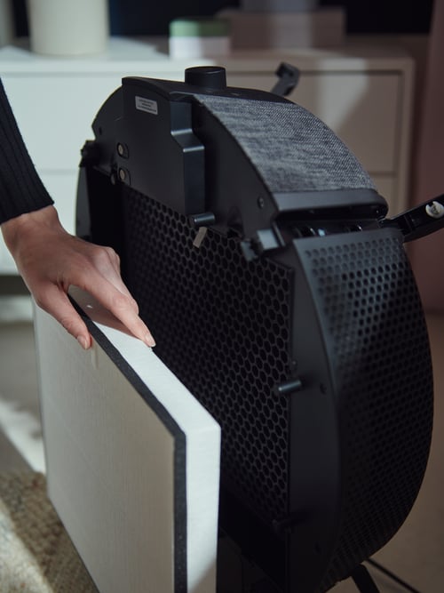 In a living room, a woman's hand removes the HEPA 12 filter from the front of a black floor model STARKVIND air purifier.
