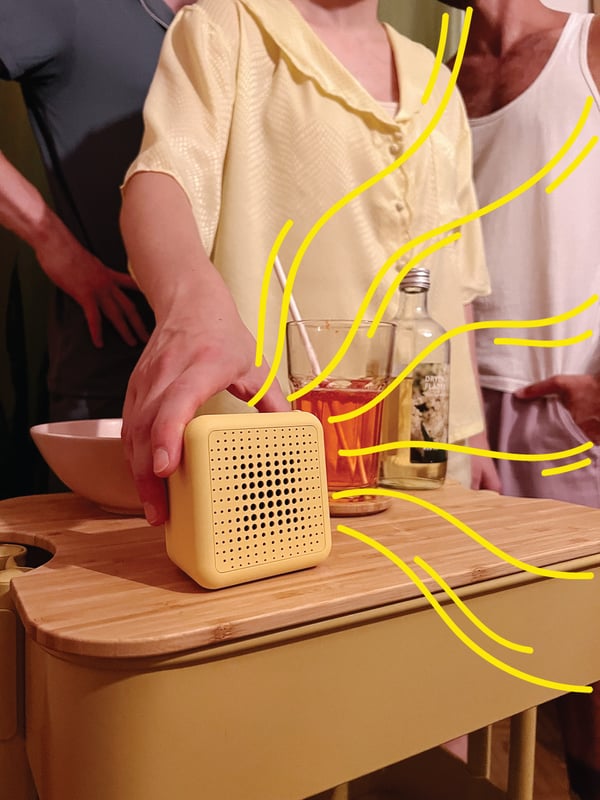 Eine Person greift nach einem gelben VAPPEBY Bluetooth-Lautsprecher, der auf einem Servierwagen steht.